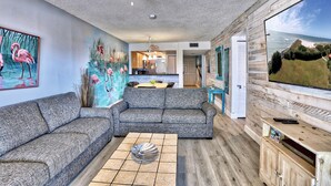 A living room with a gray sofa, a coffee table, a mural of flamingos, a ceiling fan, and a TV on a wooden wall.