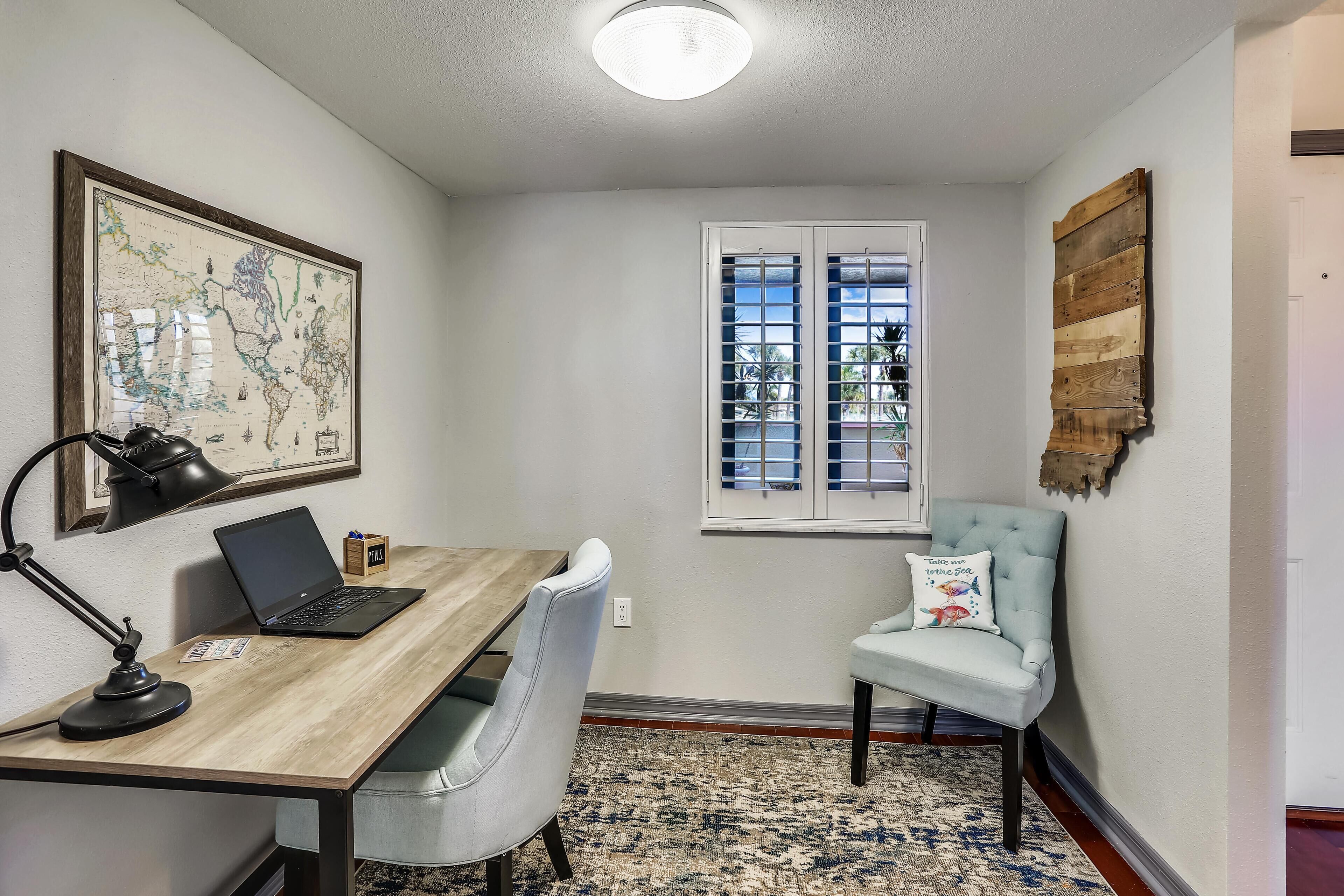 A room with a wooden desk, a laptop, a map, a window with blinds, a light blue chair, and a patterned rug.