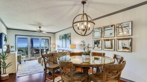 A dining area with a round table, wooden chairs, and a chandelier. There are multiple framed mirrors on the wall and a view of the outdoors through large windows. - Land's End 11-204 Beach Front (Treasure Island)