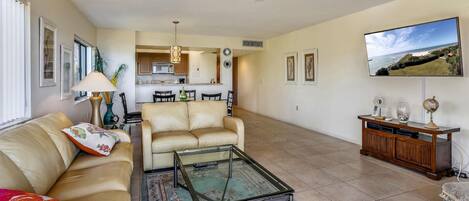 A living room with a sofa, coffee table, and TV. There is a kitchen in the background.