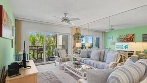 A living room with a sofa, armchairs, a coffee table, a desk with a computer, and a balcony with a view of greenery. - Land's End 11-304 Beach Front (Treasure Island)