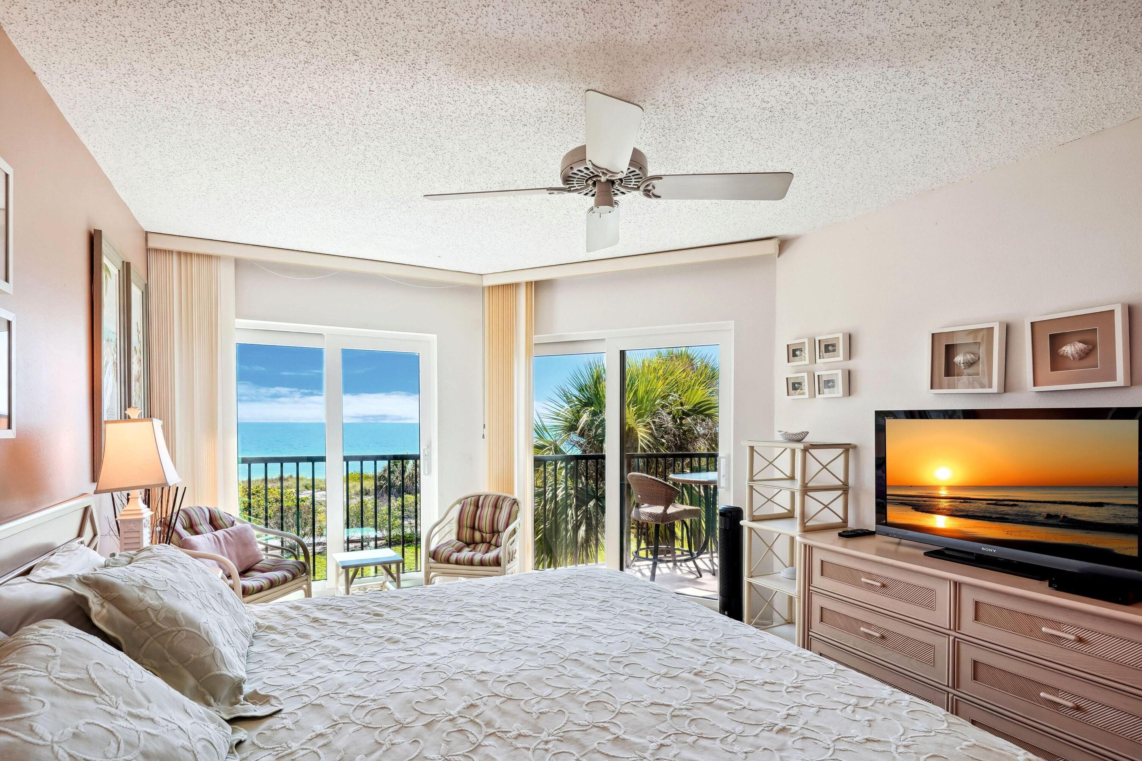 Ein Hotelzimmer mit einem großen Bett, einem Fernseher, einem Deckenventilator, einem Balkon mit Meerblick und einem Essbereich mit Stühlen.