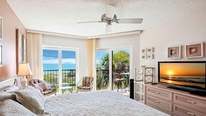 A hotel room with a large bed, a TV, a ceiling fan, a balcony with a view of the ocean, and a dining area with chairs. - Land's End 11-304 Beach Front (Treasure Island)