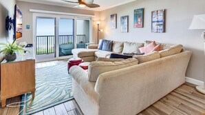 A living room with a beige sofa, a wooden cabinet, a floor lamp, and a view of the outdoors through a sliding glass door. - Land's End 11-405 Beach Front - Premier (Treasure Island)