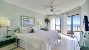 Un dormitorio con una cama grande, ventilador de techo, mesita de noche con lámpara, televisión y balcón con vista al océano.