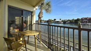 Un balcon avec une table et des chaises, donnant sur une étendue d’eau, avec un palmier et des bâtiments en arrière-plan.