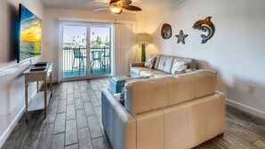 A living room with a beige sofa, a TV, a ceiling fan, and a balcony with outdoor furniture. - Land's End 3-203 Bay Front - Premier (Treasure Island)