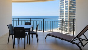 A balcony with a dining table, chairs, and a lounge chair, overlooking a clear blue sea and a tall building.