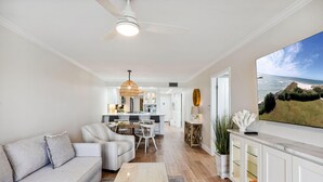 A modern living room with a sofa, coffee table, and a dining area with chairs and a table. A large TV mounted on the wall displays a scenic view. - Land's End 4-204 Bay Front - Premier (Treasure Island)