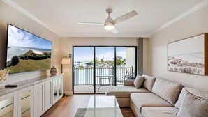 A living room with a large TV, a ceiling fan, a sofa, and a view of the ocean through the sliding glass doors. - Land's End 4-204 Bay Front - Premier (Treasure Island)