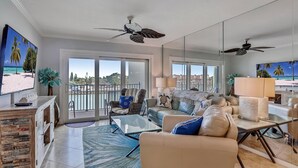 A spacious living room with a large sofa, a coffee table, and a TV. There are ceiling fans, a balcony with a view, and a large window. - Land's End 4-303 Bay Front - Premier (Treasure Island)