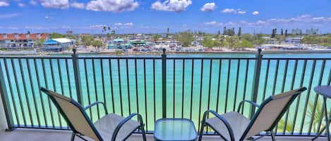 A balcony with three chairs and a table, overlooking turquoise waters and a marina with boats and buildings.