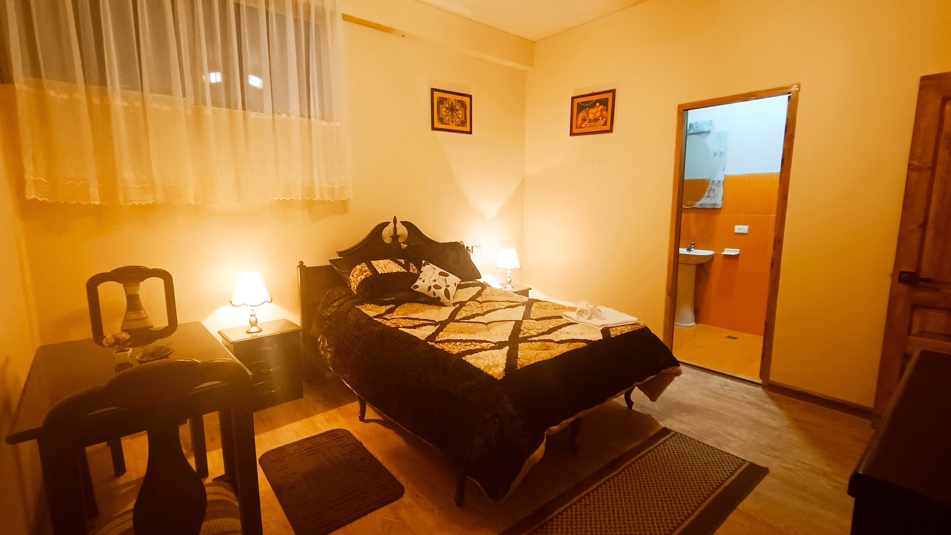 A hotel room with a bed, a chair, a table, and a nightstand. There is a window with curtains, a door, and a mirror on the wall.