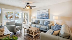 A living room with a ceiling fan, two sofas, a coffee table, and a large window with blinds. - Land's End 5-202 Gulf-bay Front - Premier (Treasure Island)