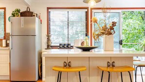 A modern kitchen with a stainless steel refrigerator, wooden bar stools, a large window with blinds, and a decorative plant on the counter.
