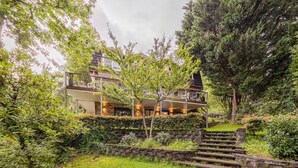 A multi-story house with a stone staircase leading to a wooden deck, surrounded by lush greenery and trees.