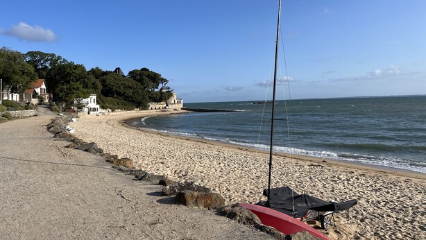 Ein Strand mit einem Segelboot, einer Steinmauer und Gebäuden im Hintergrund.