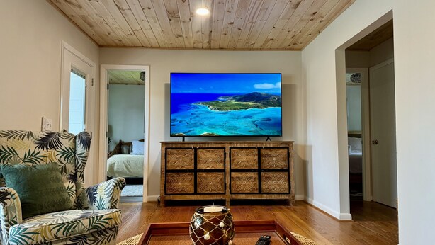 A living room with a large flat-screen TV mounted on the wall, a wooden ceiling, a patterned sofa, and a decorative tray with a candle. - Cozy cottage located in the heart of the Mayport fishing village. Pool & Patio! (Jacksonville)