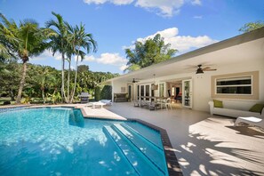 Una piscina con área de patio, mobiliario exterior y palmeras.