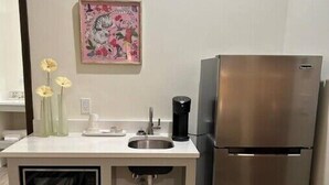 A compact kitchen with a stainless steel refrigerator, a sink, and a microwave. There is a small cabinet under the counter and a decorative painting on the wall.