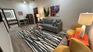 A modern living room with a grey sofa, a patterned rug, a small coffee table, and a yellow armchair. There is a painting on the wall and a lamp on a side table.