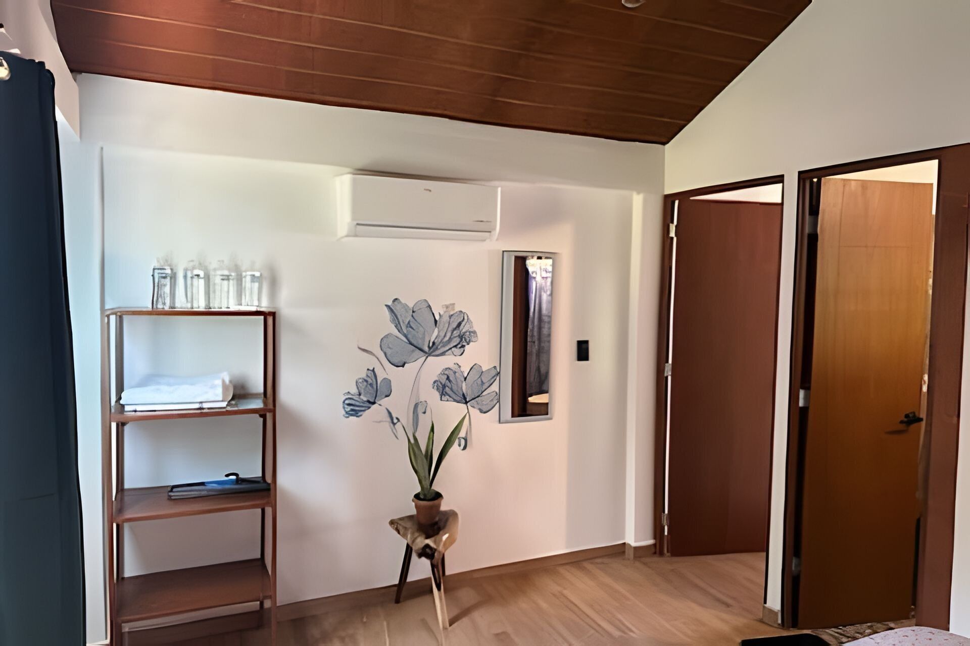A hotel room with a wooden floor, a shelf with toiletries, a wall-mounted mirror, and a door.
