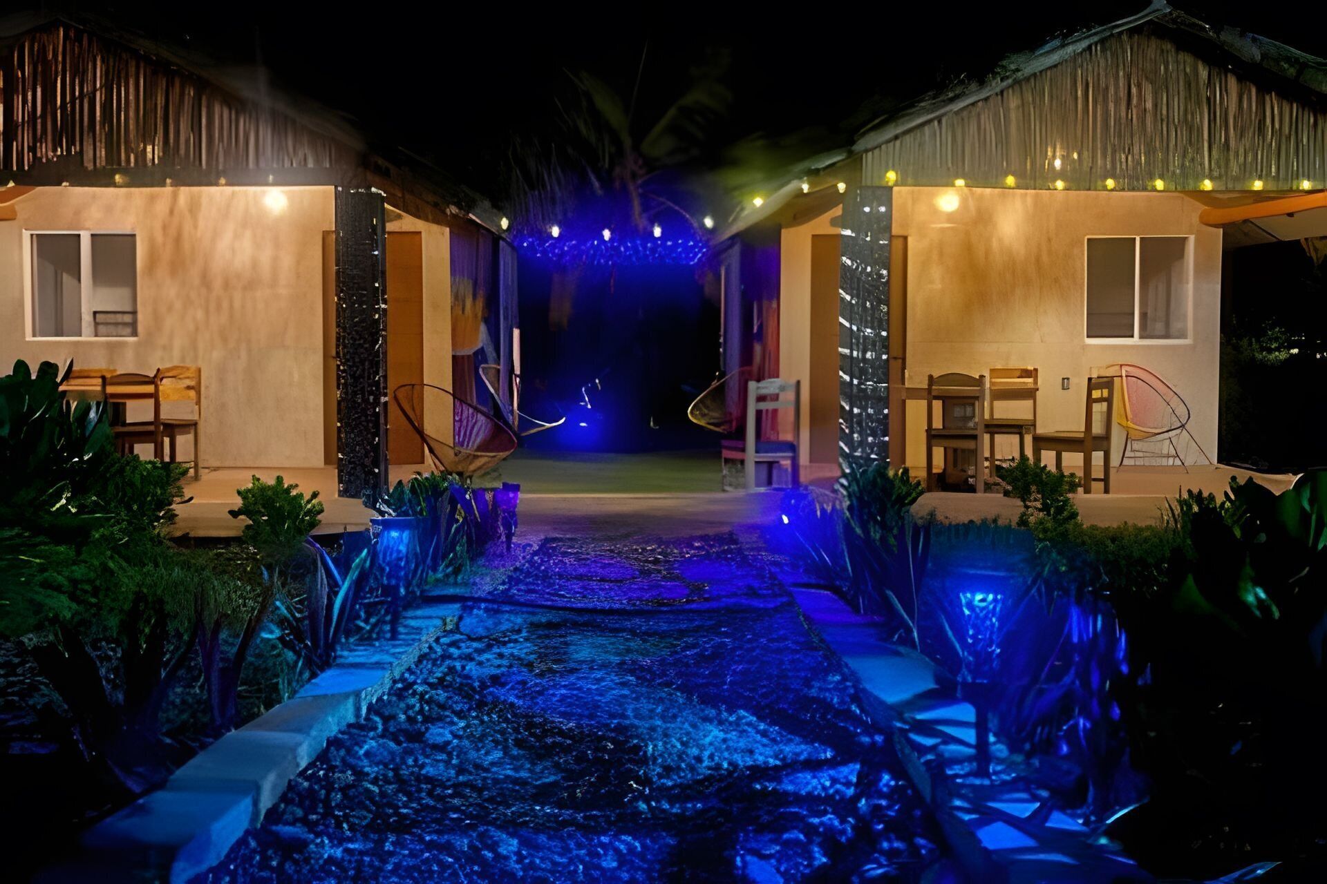 Een pad dat leidt naar een gebouw met rieten dak, omringd door stoelen en tafels, verlicht met blauw licht.