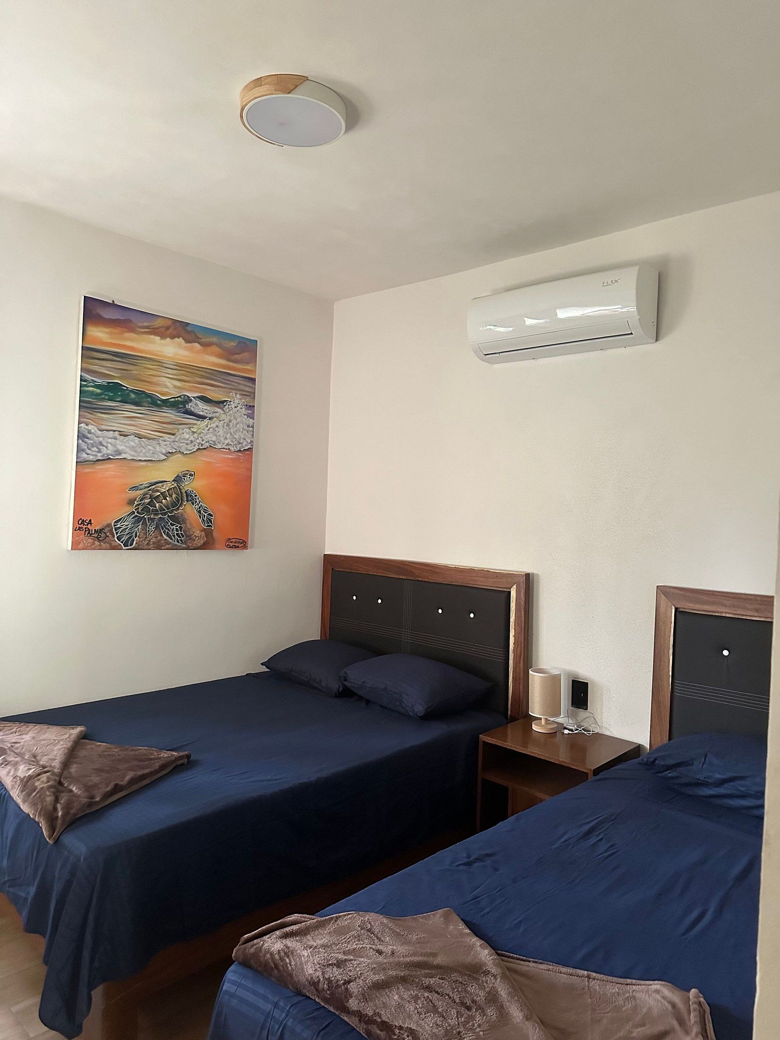 A hotel room with two single beds, a painting of a turtle on the wall, an air conditioning unit, and a bedside table with a lamp.