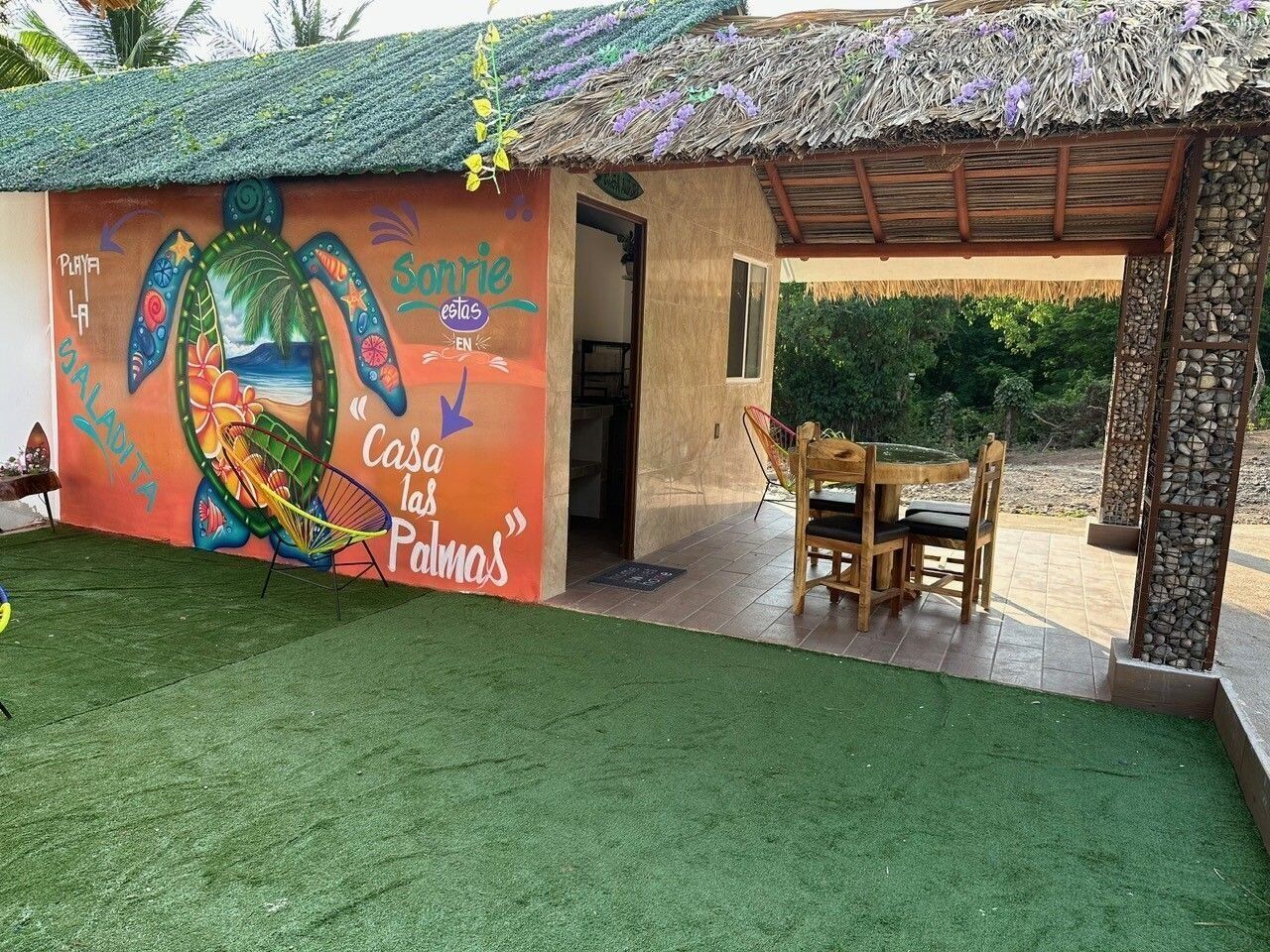 A small building with a colorful mural, a thatched roof, and a patio area with a table and chairs.