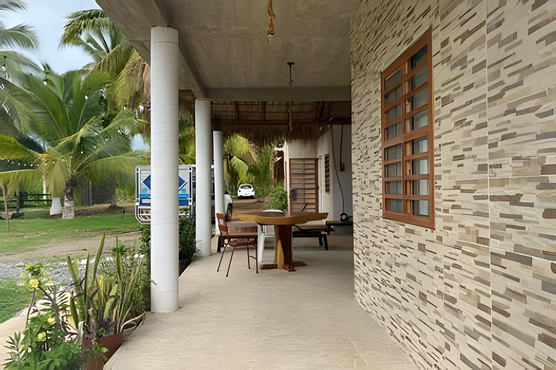 A covered outdoor area with a table and chairs, palm trees, and a building with a patterned wall and a window.
