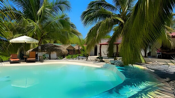 Piscina azul límpida rodeada por palmeiras e estruturas com telhados de palha, provavelmente parte de um resort.