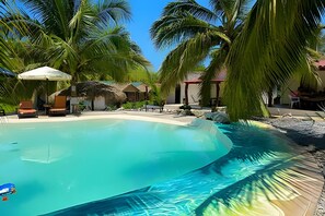 Una piscina azul cristalina rodeada de palmeras y construcciones con techo de paja, probablemente parte de un complejo turístico.