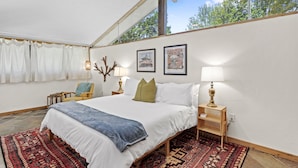 Ein Schlafzimmer mit einem großen Bett, zwei Nachttischen mit Lampen, einem Sessel und einem Fenster mit Vorhängen.