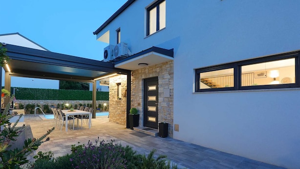 Ein modernes Haus mit Steinaußenfassade, einer überdachten Terrasse mit Gartenmöbeln und einem Schwimmbad.