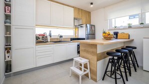 Eine moderne Küche mit weißen Schränken, einer Holztischplatte, einem Edelstahlkühlschrank und einer Obstschale. Es gibt drei schwarze Barhocker und einen kleinen weißen Hocker.