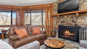A cozy living room with a stone fireplace, leather sofa, patterned ottomans, and a television mounted on a stone wall. - Cornerstone Cabin at Springdale (Haywood County)
