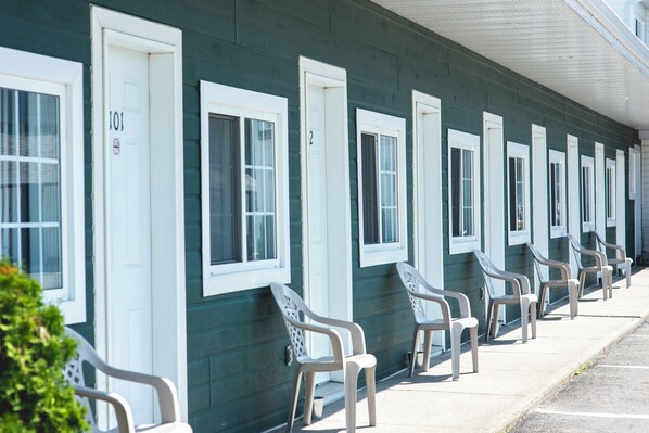 A row of motel rooms with white-framed windows and doors, numbered sequentially, and a row of plastic chairs outside.