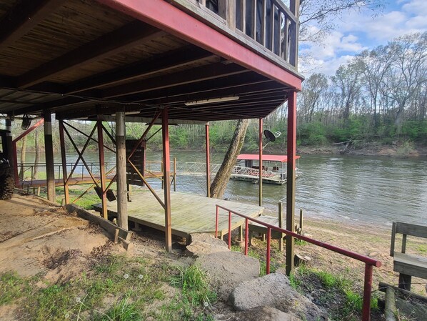 A wooden deck with a red railing overlooking a river, a dock with a boat, and a small cabin.