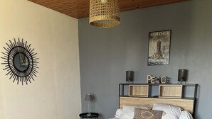 A bedroom with a wooden floor, a bed with white bedding, a hanging wicker light fixture, a wall-mounted sunburst mirror, and a framed picture of a lighthouse.
