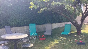 An outdoor seating area with a round table and metal chairs, surrounded by a hedge and a tree.