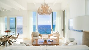 A spacious living room with a large sofa, two armchairs, a coffee table, and a chandelier. There is a glass table with chairs and a view of the ocean through large windows. - Oil Nut Bay (Virgin Gorda)