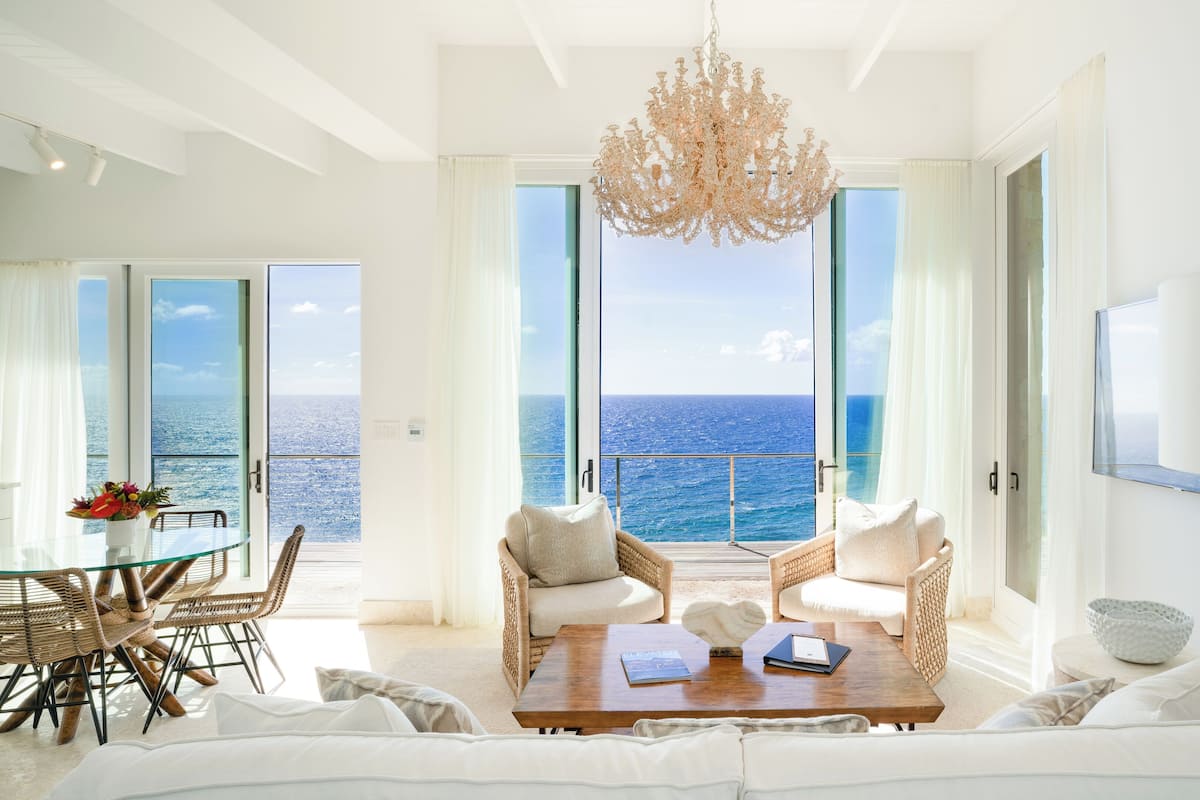 A spacious living room with a large sofa, two armchairs, a coffee table, and a chandelier. There is a glass table with chairs and a view of the ocean through large windows.