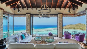 A spacious living room with a large sofa, two armchairs, and a coffee table. The room features a wooden ceiling, large windows with ocean views, and a chandelier. - Oil Nut Bay (Virgin Gorda)