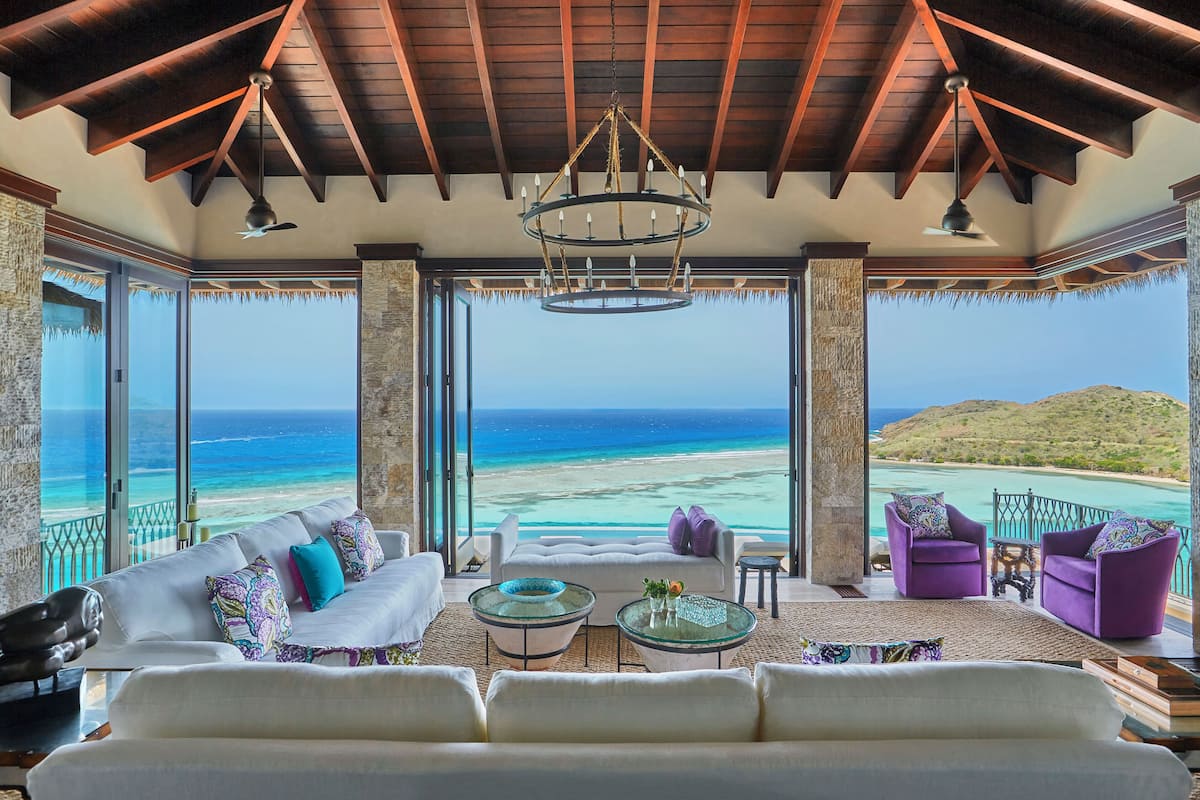 A spacious living room with a large sofa, two armchairs, and a coffee table. The room features a wooden ceiling, large windows with ocean views, and a chandelier.