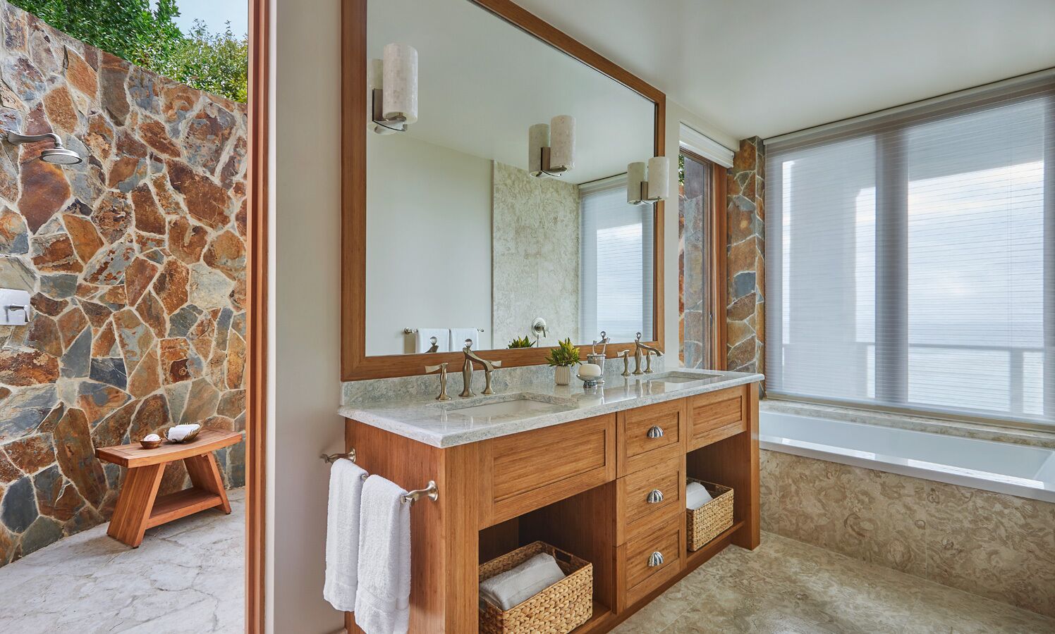 A bathroom with a stone wall, a wooden vanity with a large mirror, and a window with blinds.
