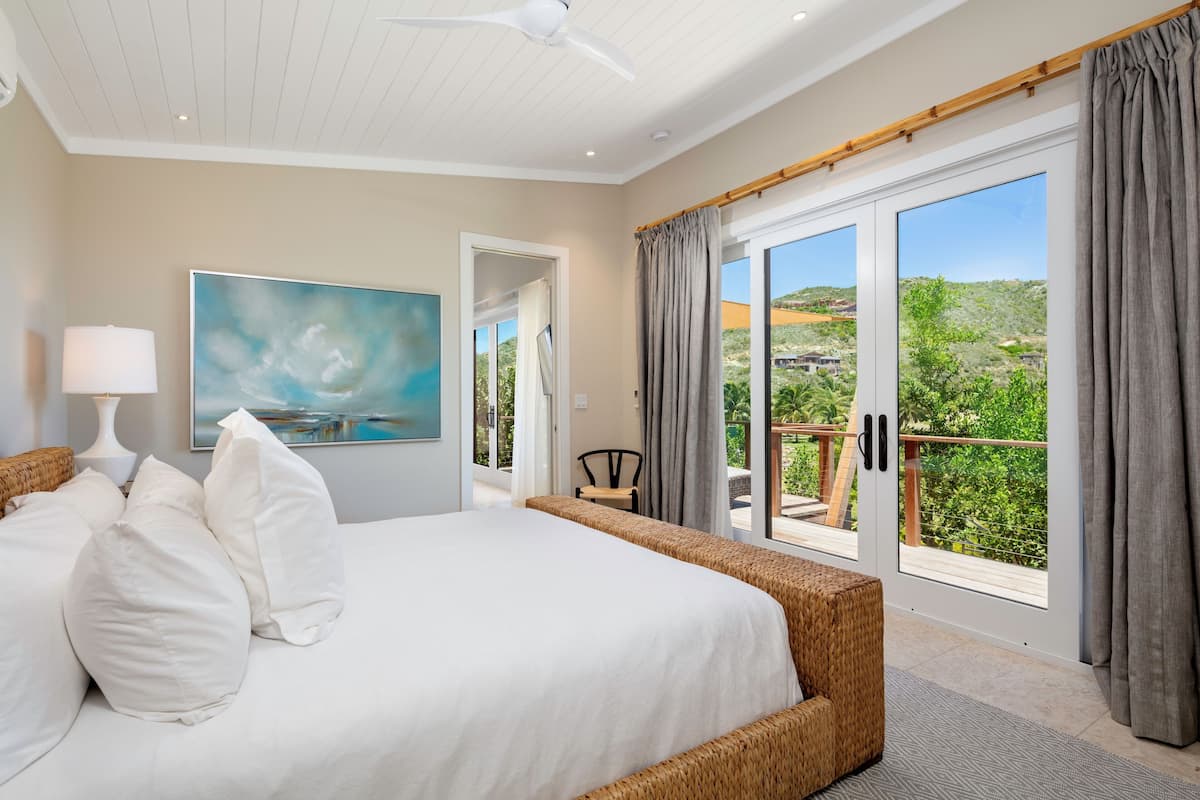A bedroom with a large bed, a ceiling fan, a painting, and a view of the outdoors.