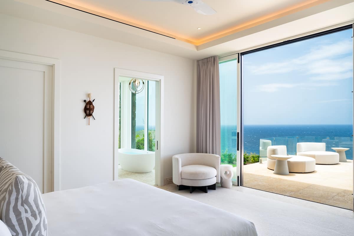 A modern bedroom with a large bed, a chair, and a view of the ocean through floor-to-ceiling glass doors.