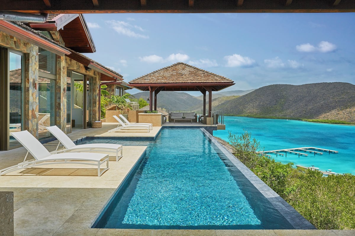 A swimming pool with lounge chairs and a gazebo overlooking a turquoise sea and mountains.