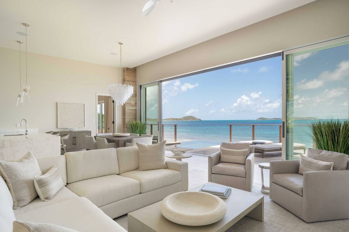 A modern living room with a large window overlooking the sea, a sofa set, and a dining area.