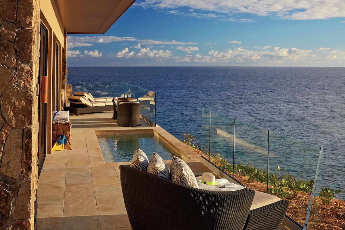A poolside area with a view of the ocean, a wicker chair, and a stone wall.